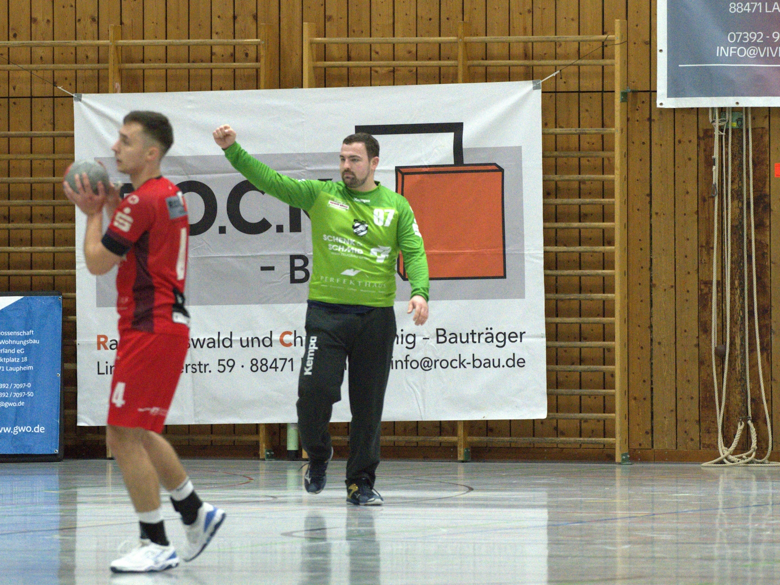 Handballspieler des HRW-Laupheim auf der linken Seite des Bildes in Ballbesitz. Im Zentrum läuft der Torwart mit erhobener Faust. Im Hintergrund sind Sponsorenplakate zu sehen